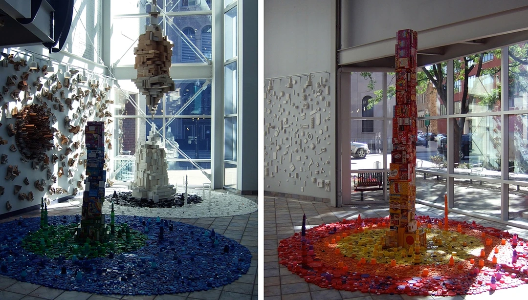 Roscoe Wilson, Beautiful Consumption IV: two views of an installation of towers built from paperboard boxes, rising from pools of plastic bottle caps in blue and green, and in red, orange and yellow, in a glass-walled atrium.