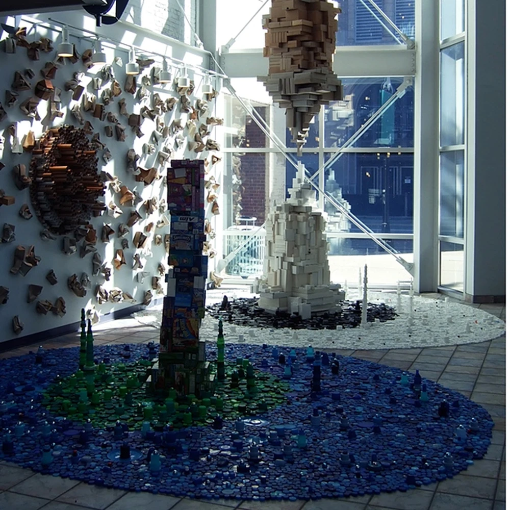 Roscoe Wilson, Beautiful Consumption IV: two views of an installation of towers built from paperboard boxes, rising from pools of plastic bottle caps in blue and green, and in red, orange and yellow, in a glass-walled atrium.