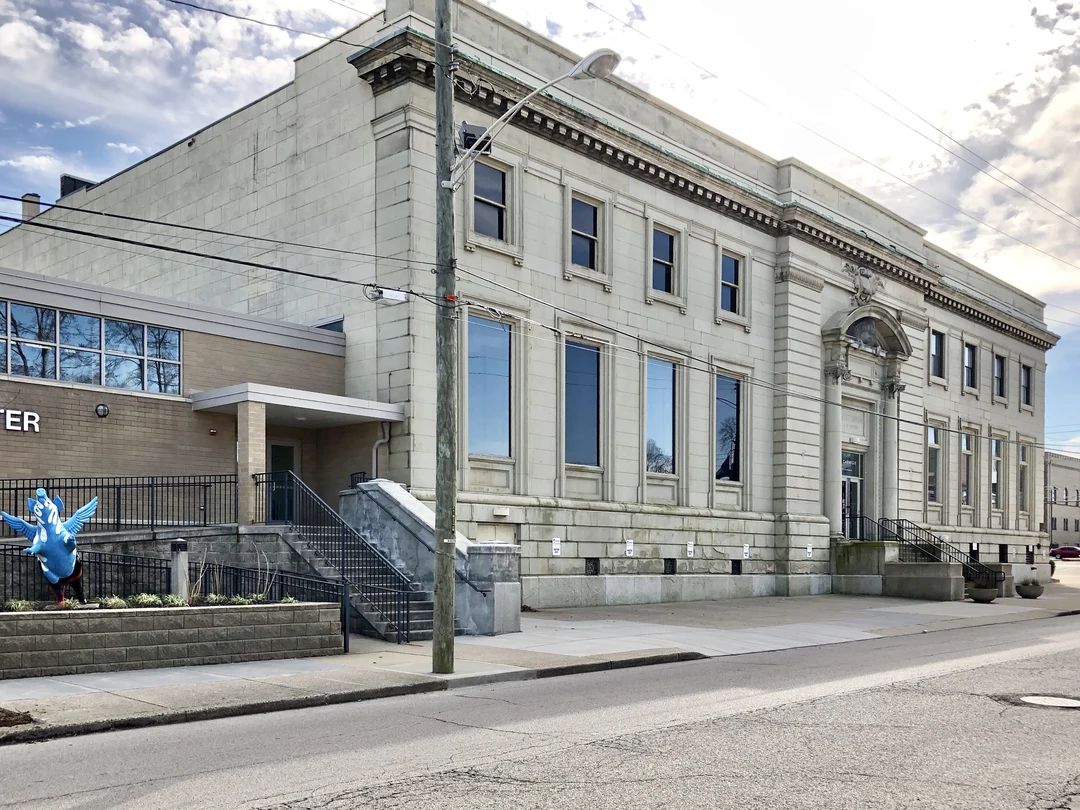 The Carnegie Visual and Performing Arts Center in Covington, Kentucky: a stone neoclassical building with tall dark windows and an arched entrance, seen from across the street with a utility pole, sidewalk, and a blue flying-pig sculpture on the left.