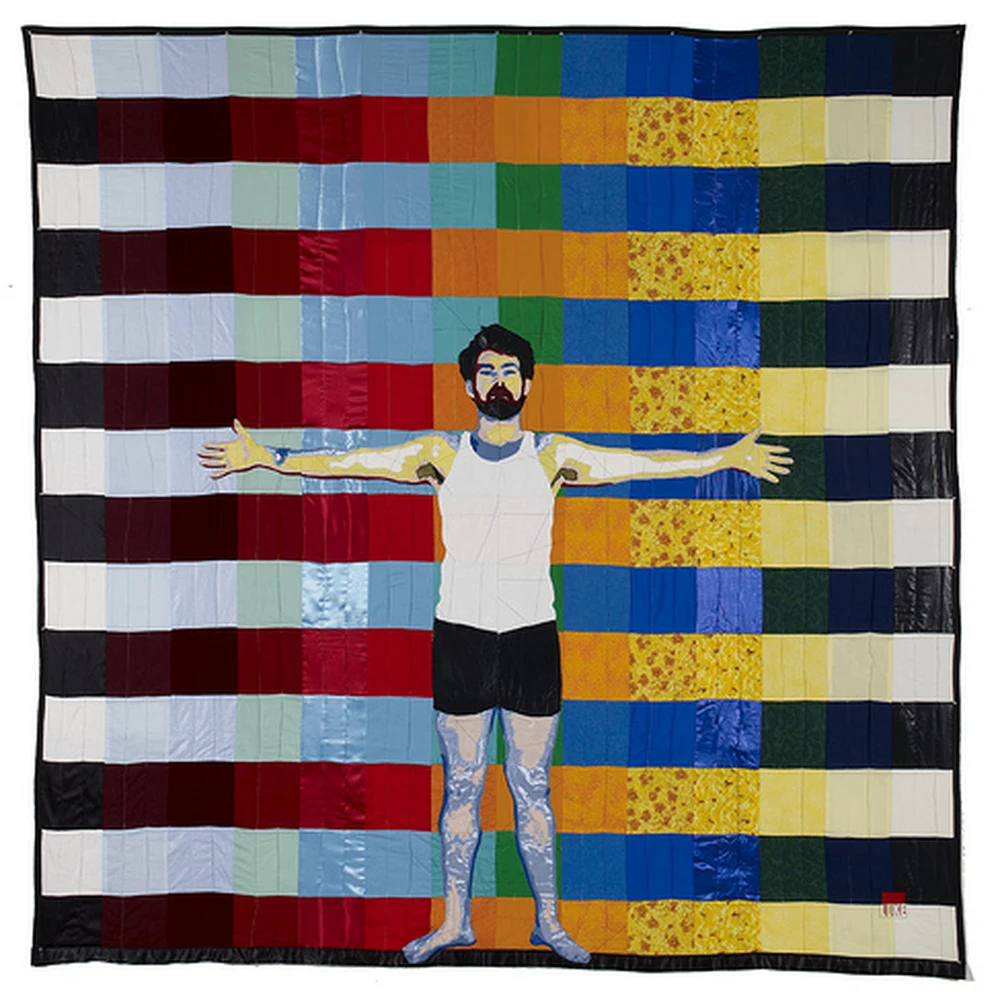 Quilted textile artwork by Luke Haynes: a bearded figure in a white tank top and dark shorts stands with arms outstretched against a colorful patchwork quilt of red, orange, yellow, green, blue and white fabric squares.