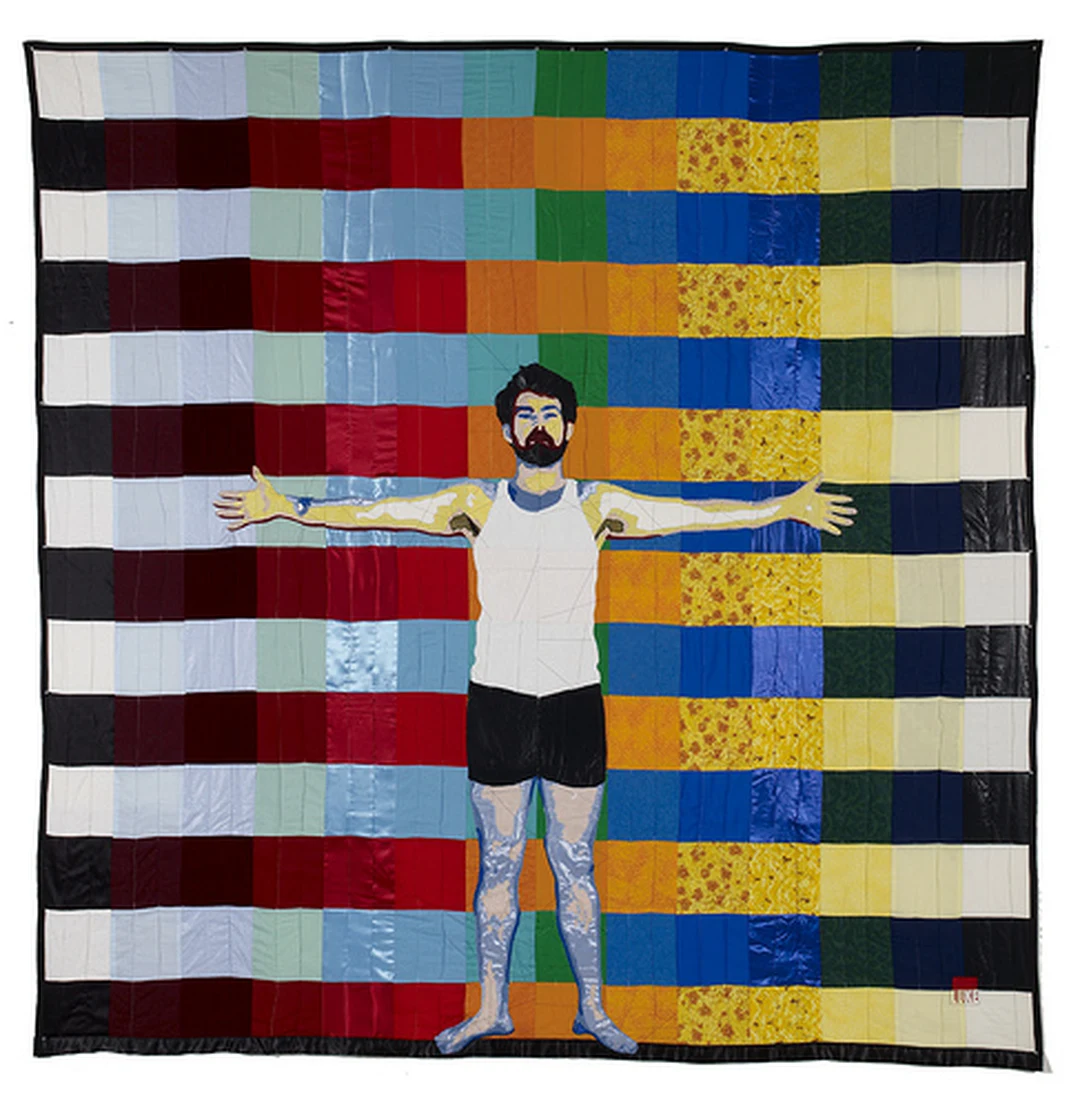 Quilted textile artwork by Luke Haynes: a bearded figure in a white tank top and dark shorts stands with arms outstretched against a colorful patchwork quilt of red, orange, yellow, green, blue and white fabric squares.