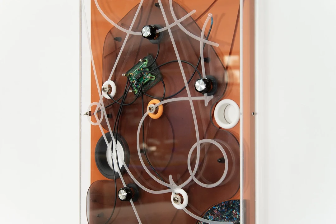 Close-up of Scott Carter's electronic sculpture Causation: a copper-toned panel set in a white frame, with a green circuit board, black and silver knobs, clear plastic tubing, black wires, and white and orange ringed ports.