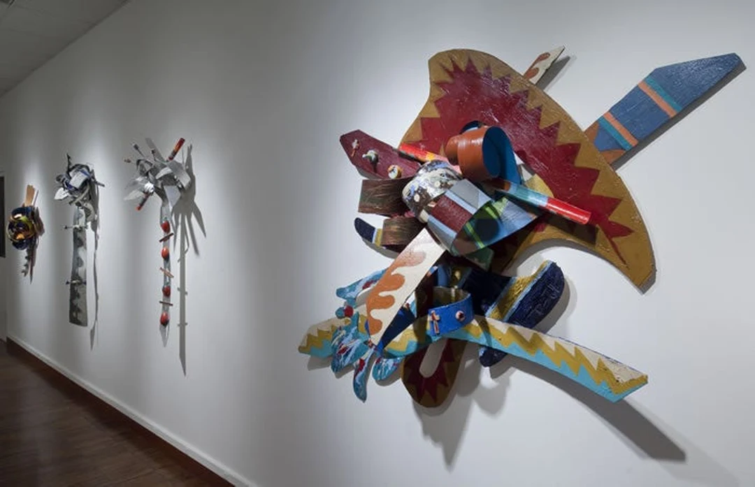 Gallery installation view: a large colorful abstract wall relief of interlocking wood forms in red, orange, blue and yellow mounted on a white gallery wall, with smaller sculptures visible further along the wall.