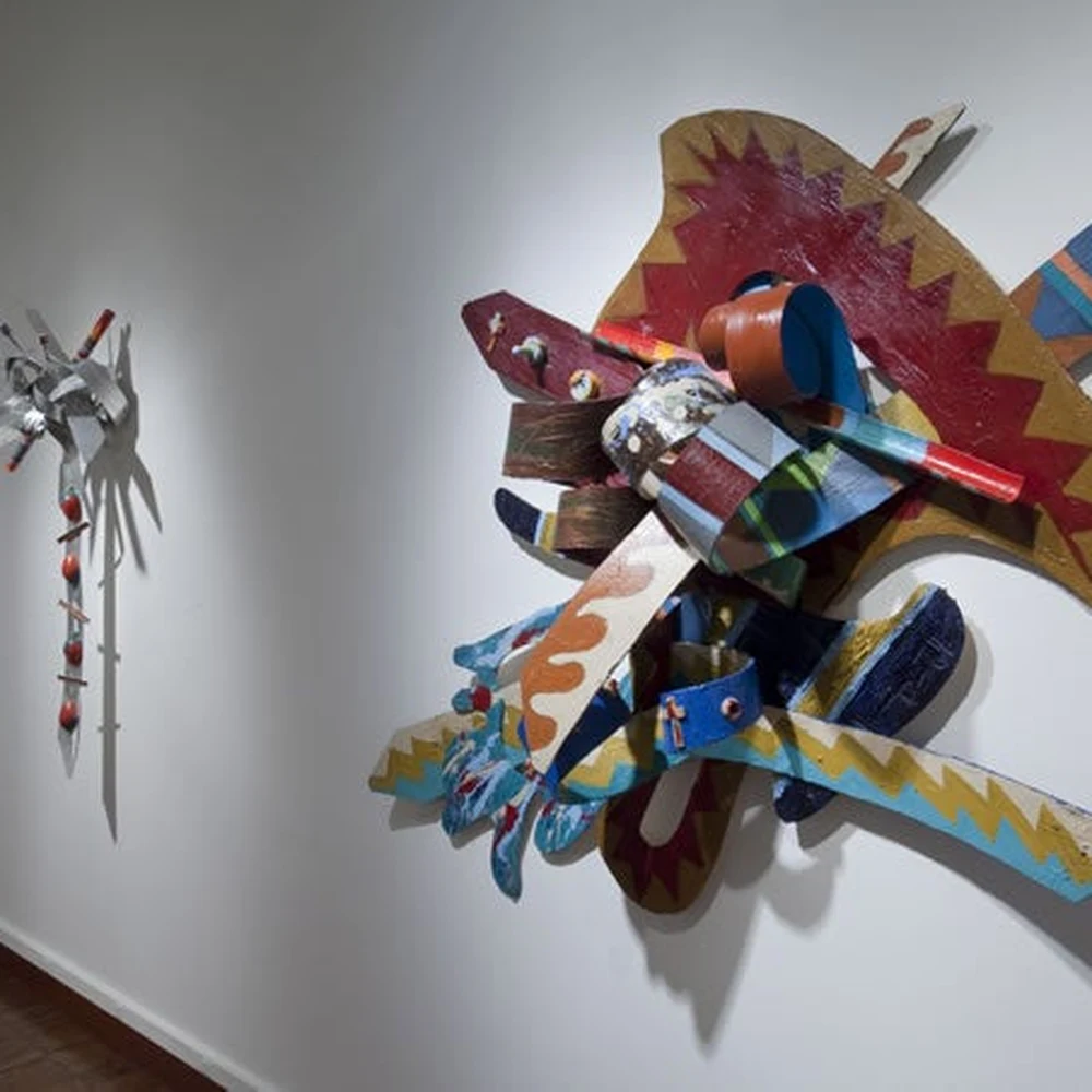Gallery installation view: a large colorful abstract wall relief of interlocking wood forms in red, orange, blue and yellow mounted on a white gallery wall, with smaller sculptures visible further along the wall.
