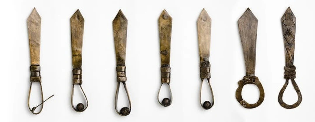 Seven cast bronze neckties hung upside down in a row like nooses against a white wall.
