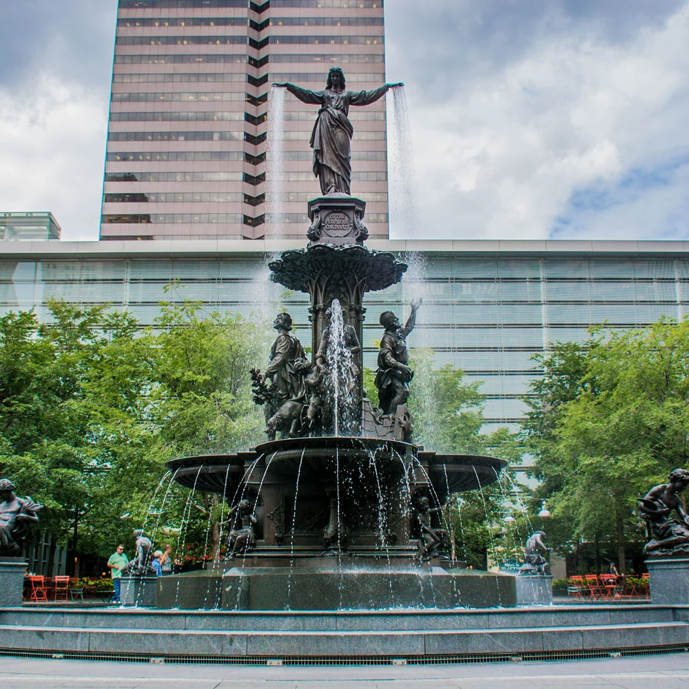The Tyler Davidson Fountain on Fountain Square, Cincinnati: a bronze fountain with a robed female figure pouring water from her outstretched hands, surrounded by smaller bronze figures, water cascading into wide basins, with trees and modern office towers behind.