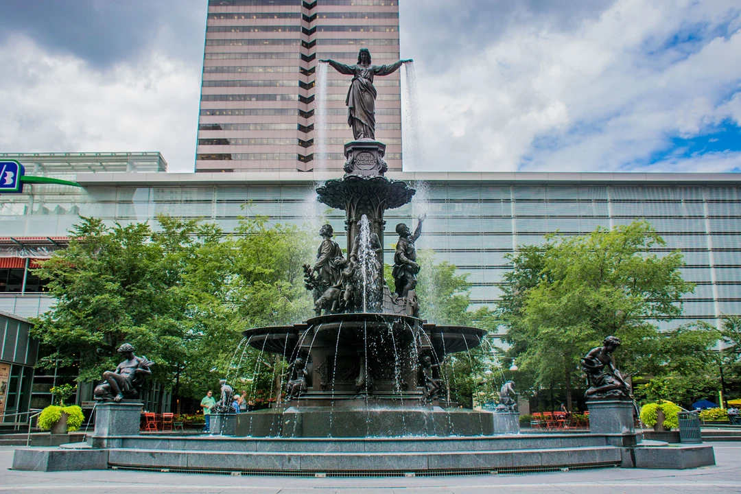 The Tyler Davidson Fountain on Fountain Square, Cincinnati: a bronze fountain with a robed female figure pouring water from her outstretched hands, surrounded by smaller bronze figures, water cascading into wide basins, with trees and modern office towers behind.