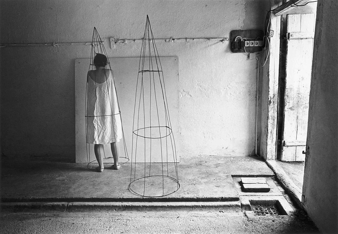 Black-and-white self-portrait by Anne Arden McDonald: the artist, seen from behind in a white dress, stands beside two tall wire cone structures in a bare abandoned room with a weathered wooden door, concrete floor and an old electrical panel on the wall.