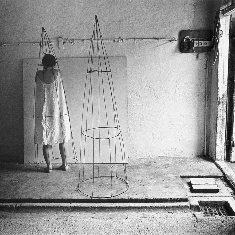 Black-and-white self-portrait by Anne Arden McDonald: the artist, seen from behind in a white dress, stands beside two tall wire cone structures in a bare abandoned room with a weathered wooden door, concrete floor and an old electrical panel on the wall.