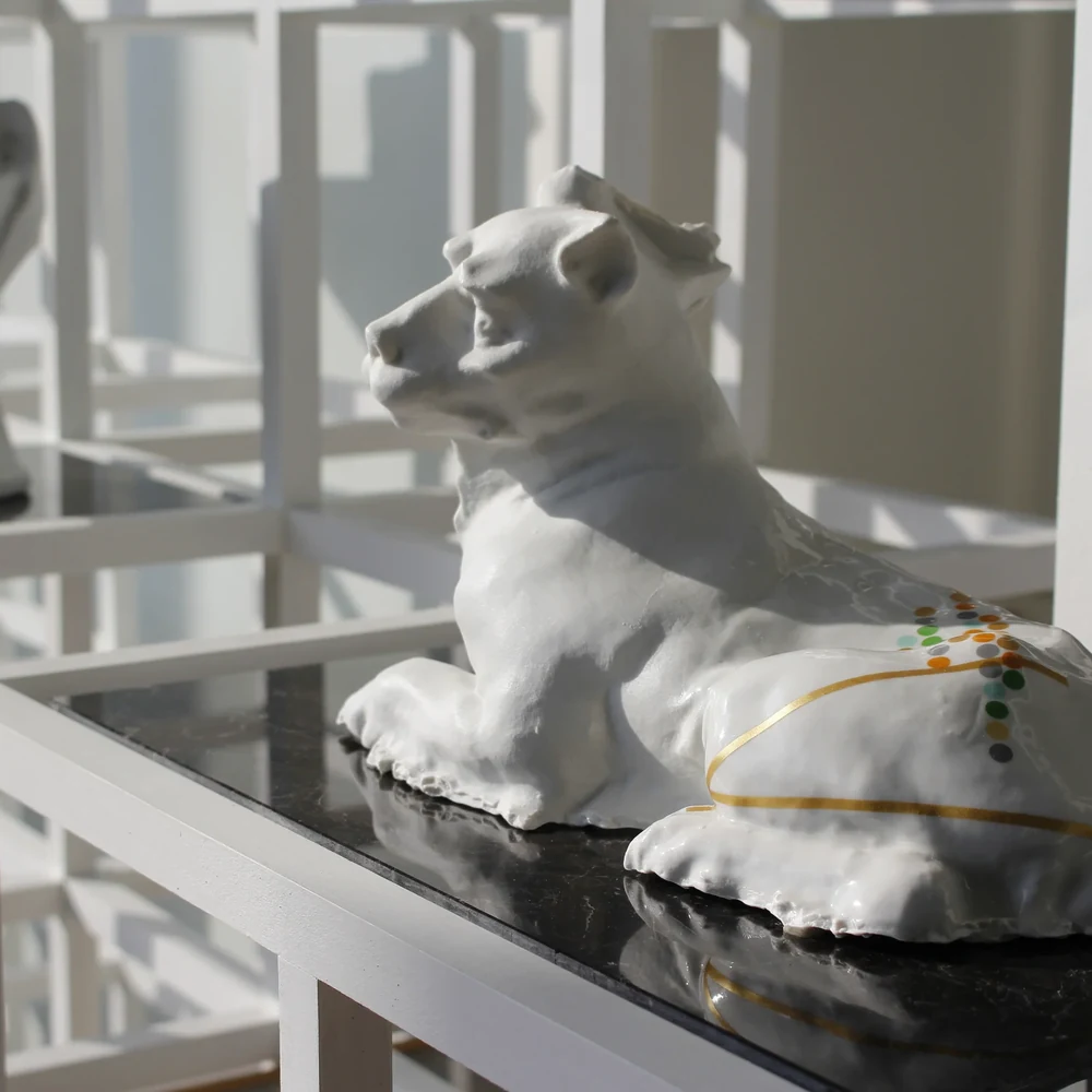 White ceramic sculpture of a reclining lioness in left profile, its haunch painted with thin gold lines and a cluster of small multicolored dots, resting on a glossy black shelf.