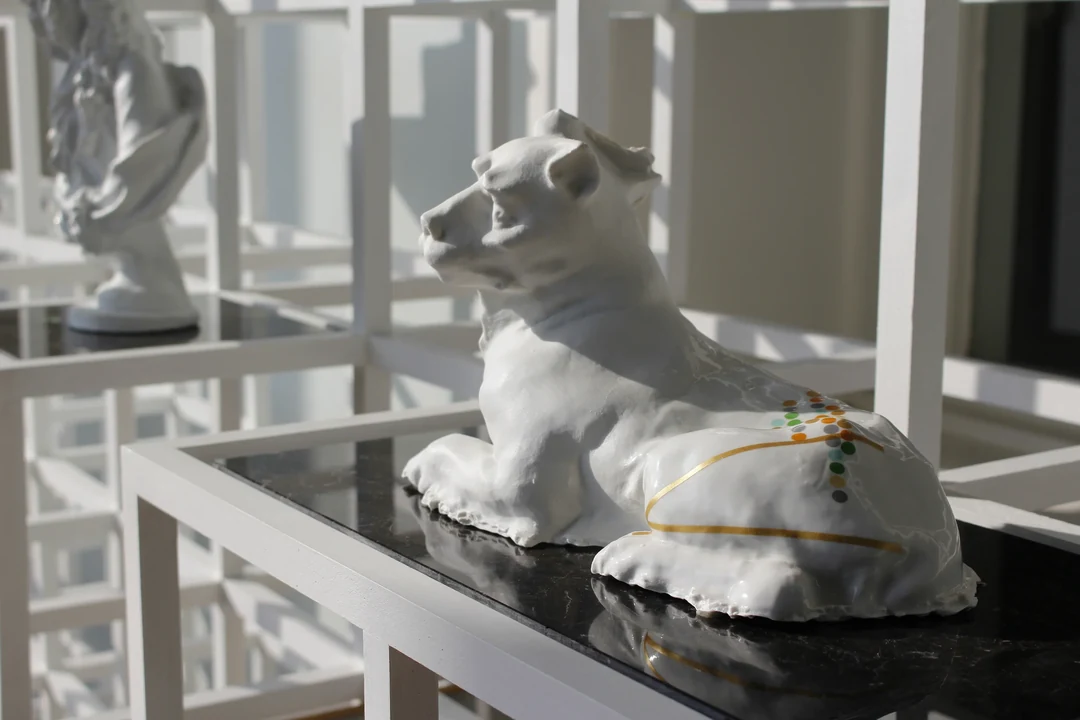 White ceramic sculpture of a reclining lioness in left profile, its haunch painted with thin gold lines and a cluster of small multicolored dots, resting on a glossy black shelf; behind it, a white grid of open cubic frames with a second white sculpted bust softly out of focus.