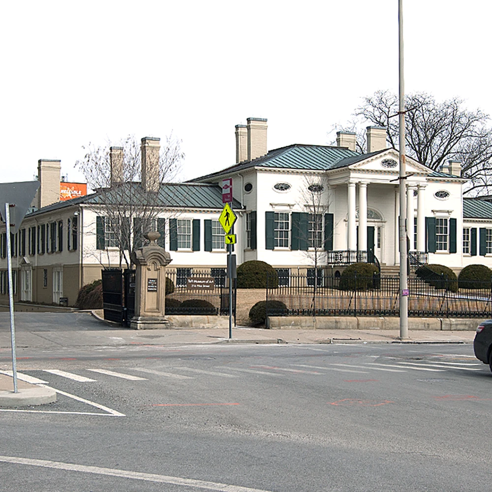 Street view of the Taft Museum of Art in Cincinnati: the white neoclassical mansion with tall columns stands behind a black iron fence, seen across the street with a stone gatepost, crosswalk lines, a pedestrian sign and a parked car.