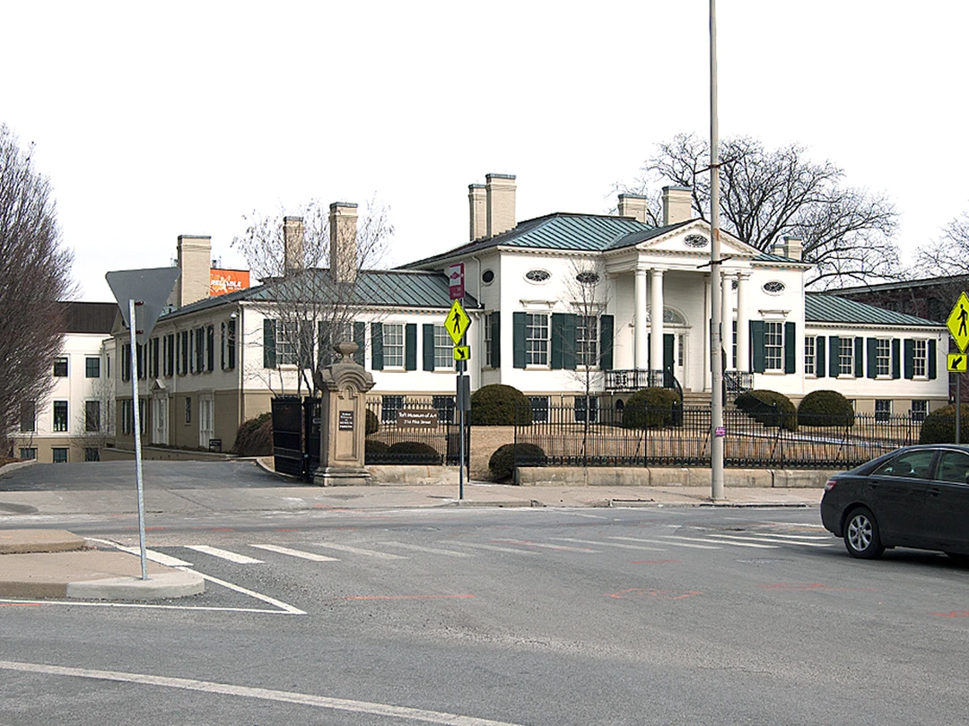 Street view of the Taft Museum of Art in Cincinnati: the white neoclassical mansion with tall columns stands behind a black iron fence, seen across the street with a stone gatepost, crosswalk lines, a pedestrian sign and a parked car.