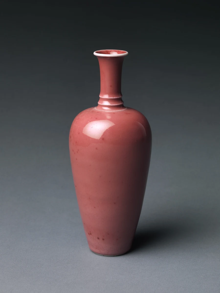 Tall bottle-shaped vase with a long neck, covered in a mottled pale copper-red peach-bloom glaze with darker speckling, photographed against a dark background. Chinese, Kangxi period (1662–1722), Bottle vase with peach-bloom glaze, porcelain, H. 7 3/4 in. (19.7 cm). Image from The Metropolitan Museum of Art. (Related image located during this archive's reconstruction.)