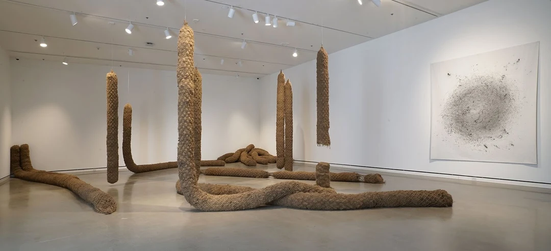 Tall brown woven-fiber columns hang from a white gallery ceiling and lie across the floor, next to a large white wall drawing with a dark circular vortex; installation view of Alexa Horochowski’s work.
