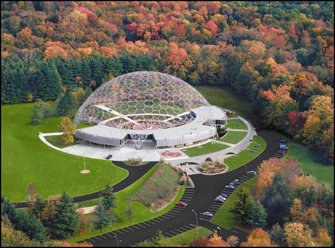 Aerial view of the ASM International headquarters near Cleveland: a vast open geodesic dome of metal latticework over a curved modernist building, set on green lawns amid autumn forest.
