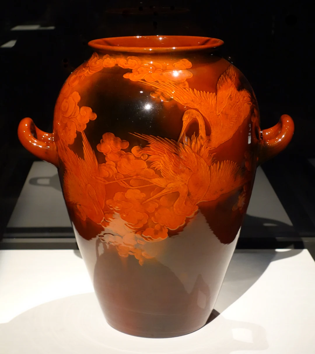 Glossy Rookwood earthenware vase with deep red-brown mahogany glaze, decorated with orange cranes flying among stylized clouds in Japonesque style, photographed in a museum display.