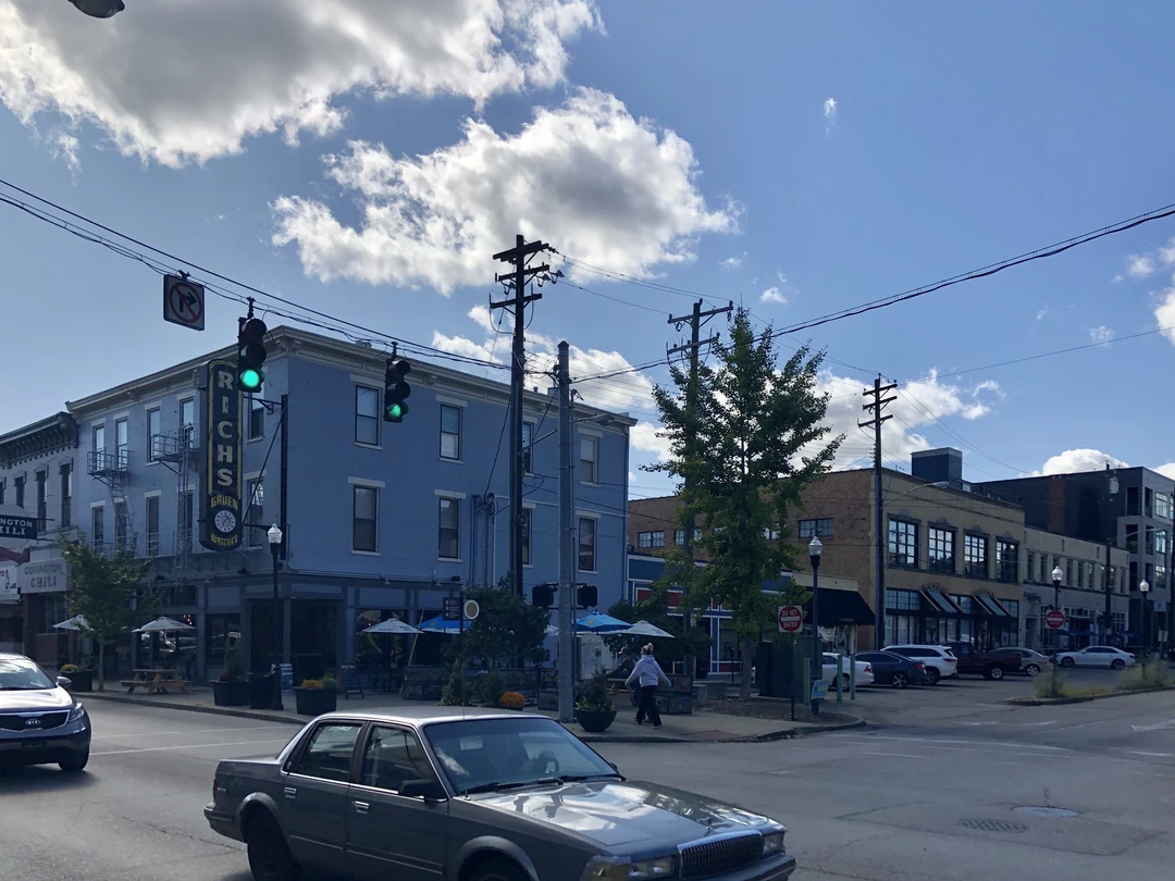 Street corner at Madison Avenue and 7th Street in Covington, Kentucky: a blue two-story brick building with a vertical sign, sidewalk umbrellas and traffic lights under a bright cloudy sky.