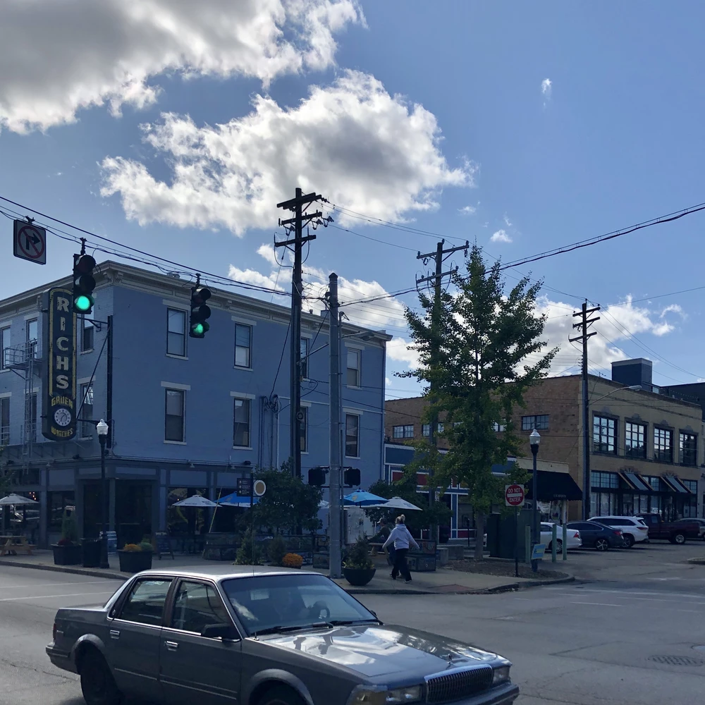 Street corner at Madison Avenue and 7th Street in Covington, Kentucky: a blue two-story brick building with a vertical sign, sidewalk umbrellas and traffic lights under a bright cloudy sky.