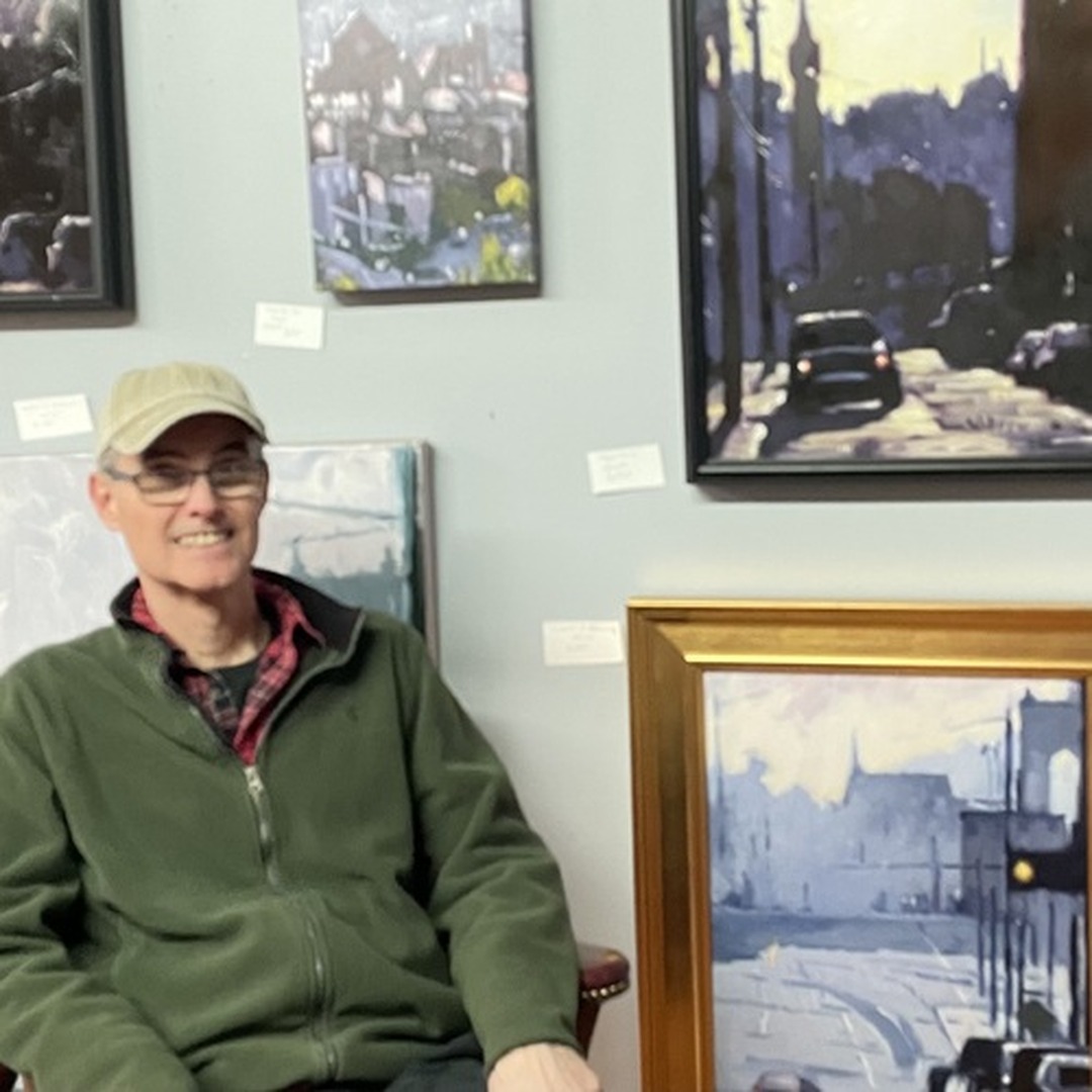 Oil painter Paul Wolven in a gallery, with his paintings hanging on the wall behind him.