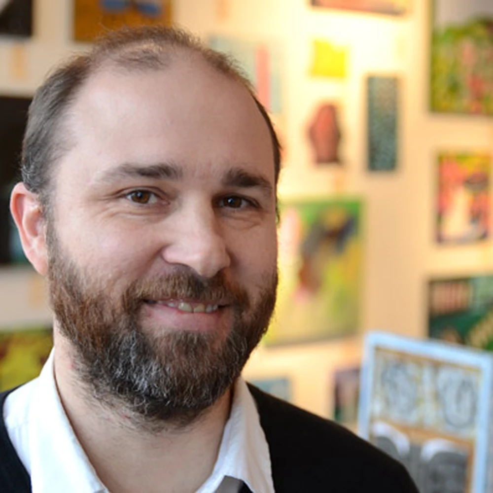 Close-up portrait of Matt Distel, a bearded man in a black sweater and white shirt, smiling, with colorful artworks on a gallery wall behind him.