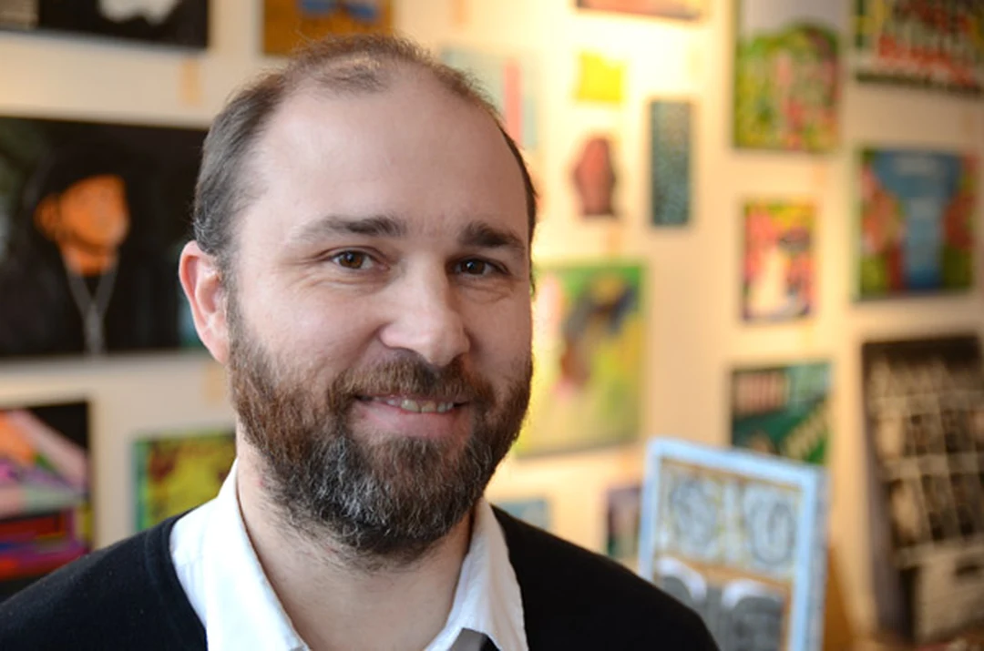 Close-up portrait of Matt Distel, a bearded man in a black sweater and white shirt, smiling, with colorful artworks on a gallery wall behind him.