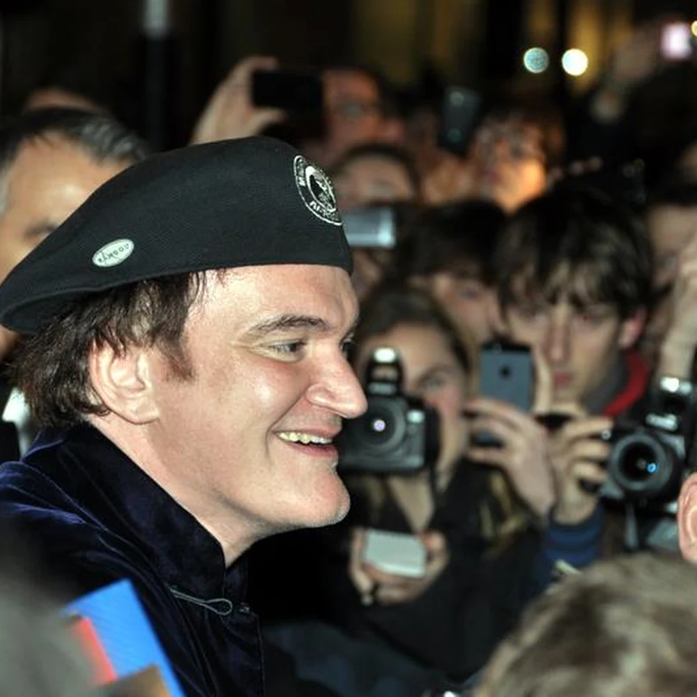 Quentin Tarantino wearing a black beret, smiling amid a crowd of photographers at a film premiere.