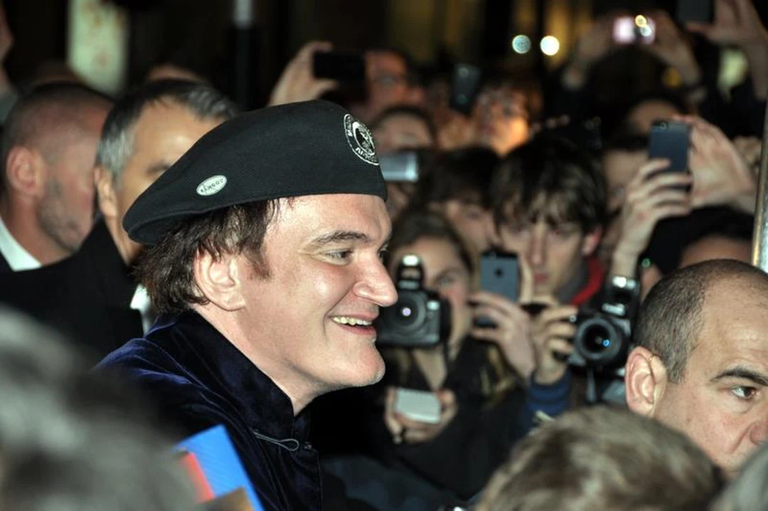 Quentin Tarantino wearing a black beret, smiling amid a crowd of photographers at a film premiere.