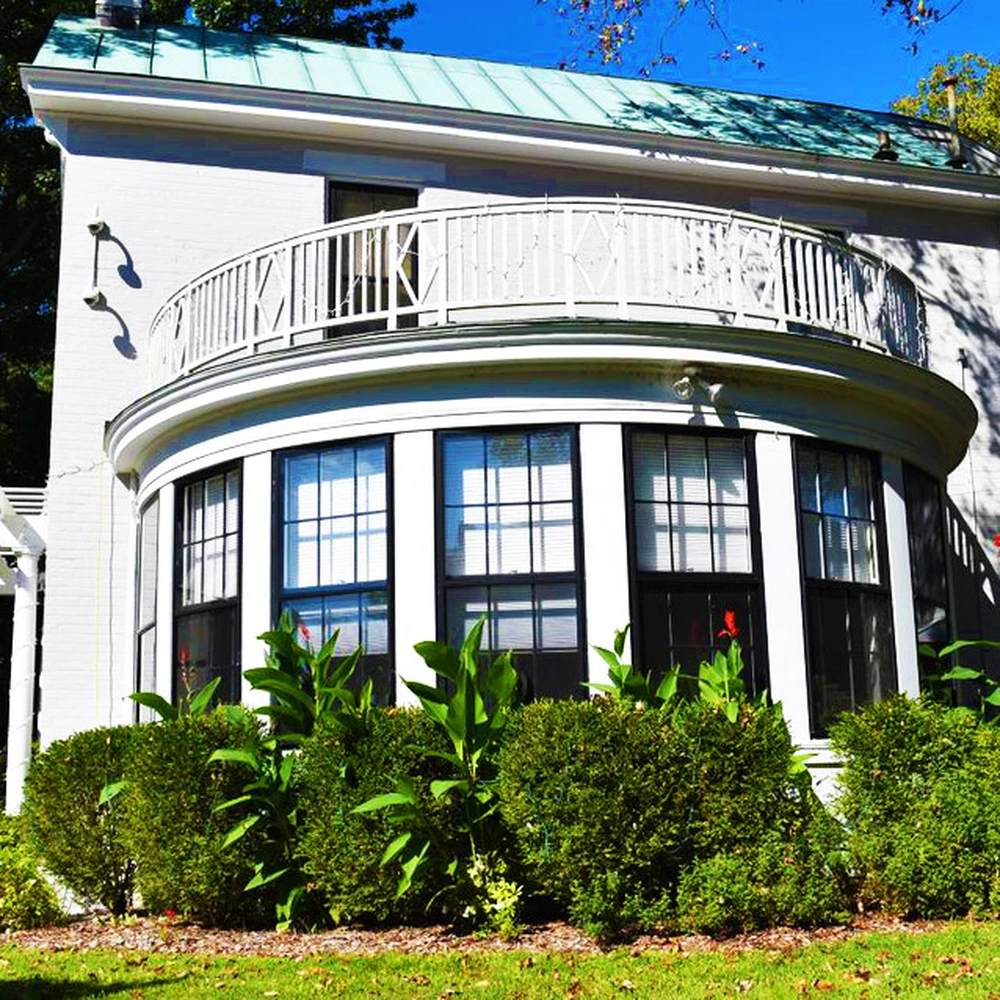 White two-story building with a curved second-floor balcony and tall windows, set among trees and garden plantings under a blue sky.