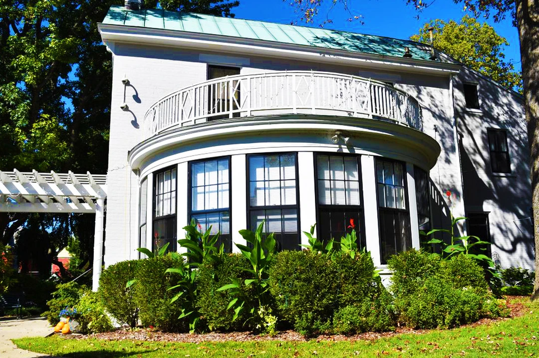 White two-story building with a curved second-floor balcony and tall windows, set among trees and garden plantings under a blue sky.