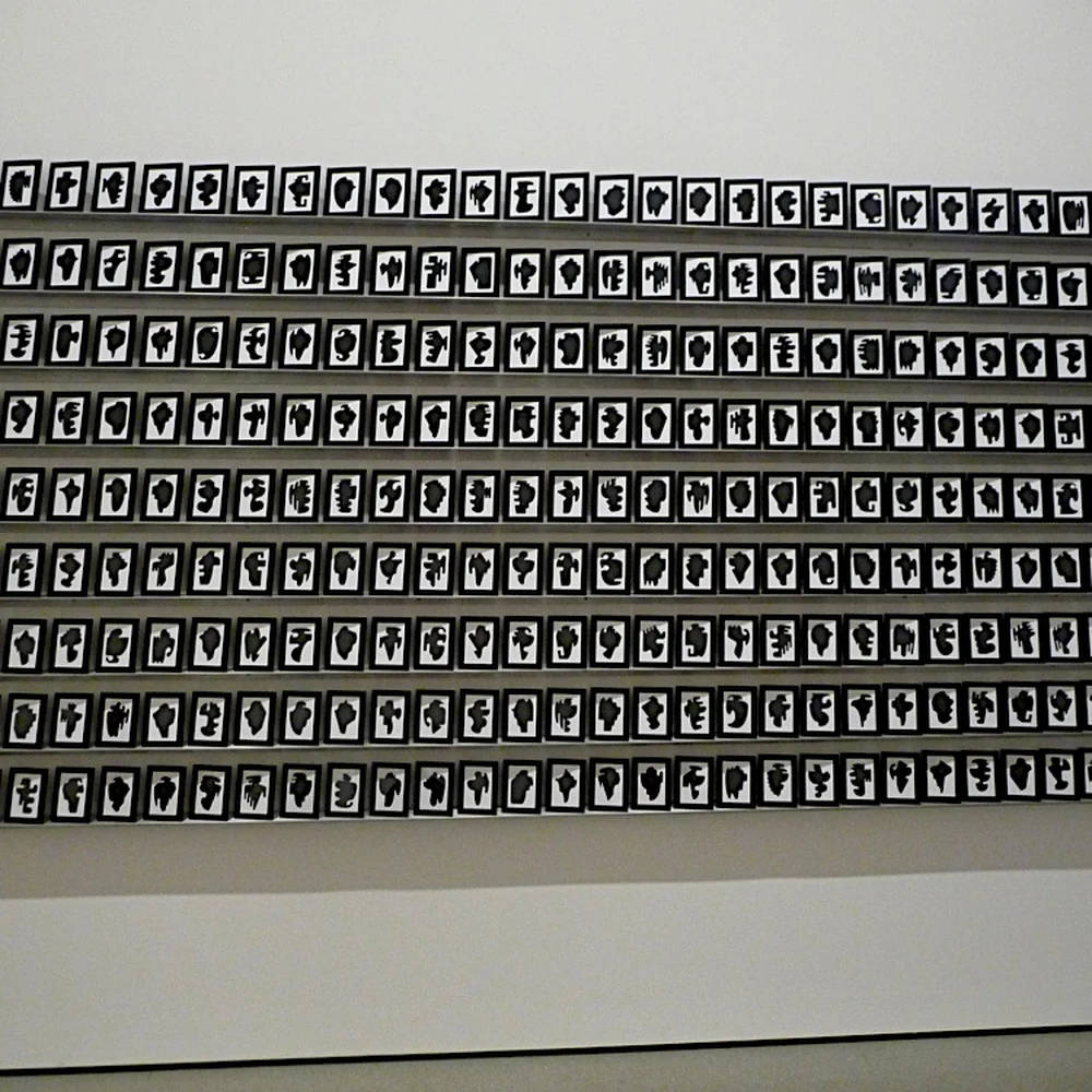 Installation view of Allan McCollum's The Shapes Project: hundreds of small framed black shapes on white paper covering a museum wall in a tight grid.