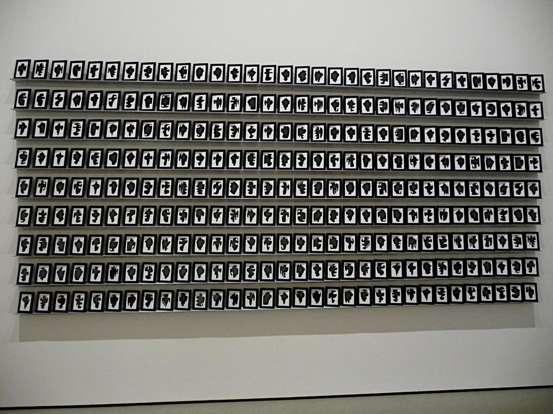 Installation view of Allan McCollum's The Shapes Project: hundreds of small framed black shapes on white paper covering a museum wall in a tight grid.