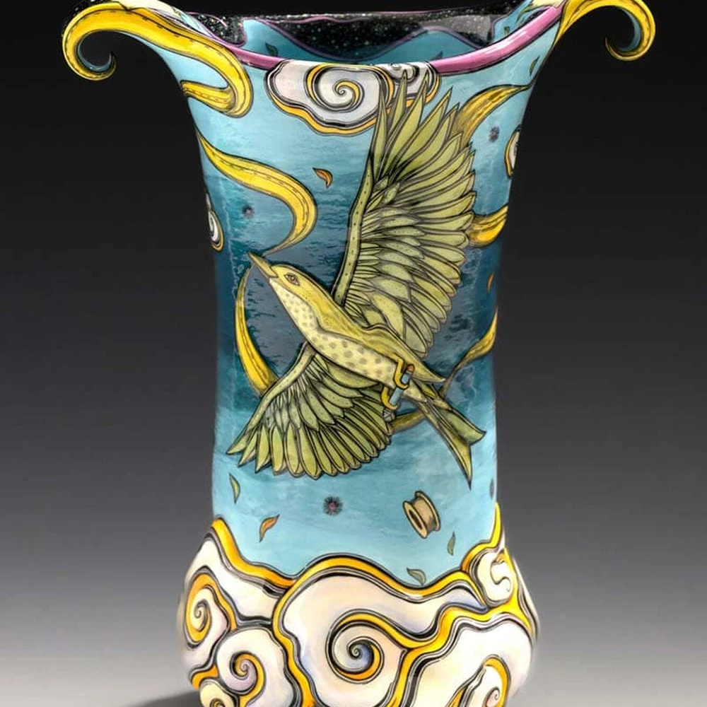 A ceramic vessel by Terri Kern, painted with a bird in flight among stylized clouds in blue, yellow and white.