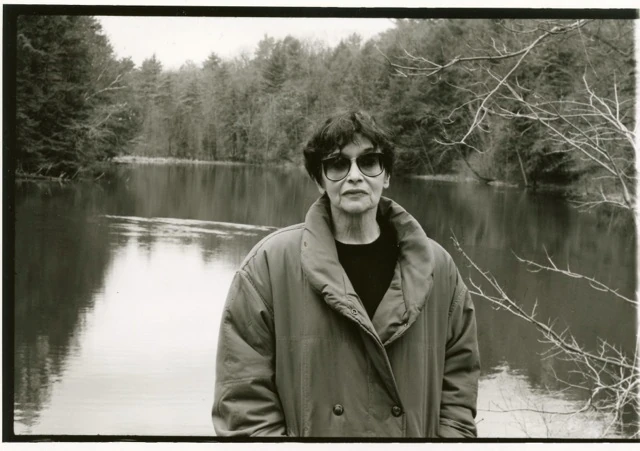 Black-and-white photograph of writer Carole Klein standing outdoors before a calm lake, wearing sunglasses and a puffy winter coat, trees and their reflections behind her.. Louis Zoeller Bickett, Carole Klein at Yaddo, 1995. Image from the AEQAI archive.