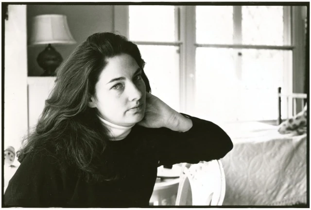 Black-and-white portrait of writer A.M. Homes, hand resting at her neck, long dark hair, dark turtleneck; a lamp and a sunlit window with a bed behind her.. Louis Zoeller Bickett, A.M. Homes at Yaddo, 1995. Image from the AEQAI archive.