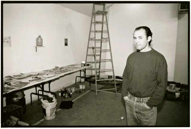 Black-and-white photograph of artist Andrew Castrucci standing in his studio at Yaddo, hands in pockets; behind him a long worktable covered with prints, a tall stepladder, a jug on a wall shelf and a paint bucket on the floor.. Louis Zoeller Bickett, Andrew Castrucci in His Studio at Yaddo, 1995. Image from the AEQAI archive.
