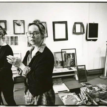 Black-and-white photograph of the identical Kenyon twins standing in their studio at Yaddo; the twin in the foreground wears glasses and clasps her hands, framed artworks hang on the wall behind them, a floor lamp and a white side table to the right.