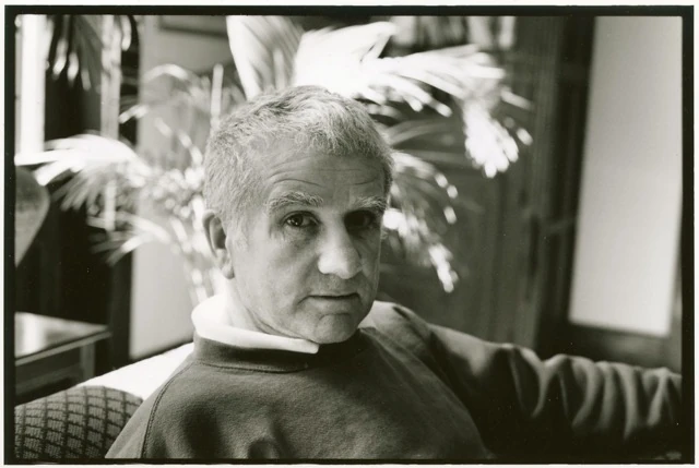 Black-and-white portrait of writer Joseph Caldwell seated indoors, short gray hair, wearing a dark sweatshirt over a white turtleneck, palm fronds behind him, one arm resting on the chair.. Louis Zoeller Bickett, Joseph Caldwell at Yaddo, 1995. Image from the AEQAI archive.