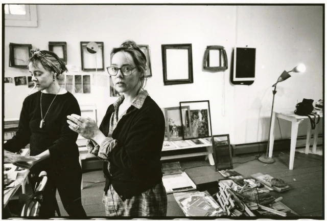 Black-and-white photograph of the identical Kenyon twins standing in their studio at Yaddo; the twin in the foreground wears glasses and clasps her hands, framed artworks hang on the wall behind them, a floor lamp and a white side table to the right.