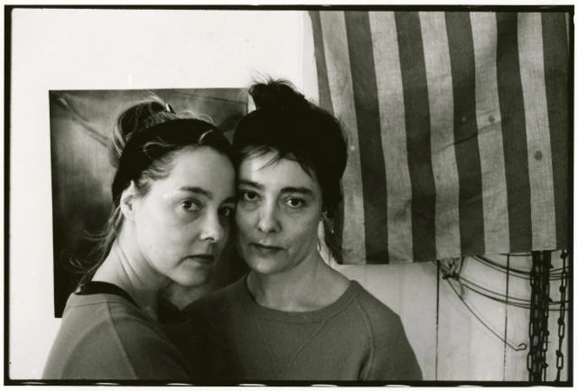 Black-and-white close portrait of the identical Kenyon twins side by side in their studio; a striped textile and loose hanging wires on the wall behind them.. Louis Zoeller Bickett, The Kenyon Twins in Their Studio at Yaddo (1), 1995. Image from the AEQAI archive.