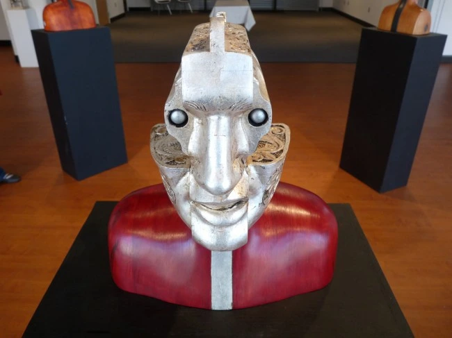 Wood and metal sculpture of a head and shoulders whose face is the ornate silver cylinder of a revolver, with bullets for eyes.