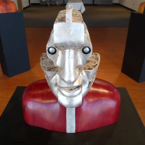 Wood and metal sculpture of a head and shoulders whose face is the ornate silver cylinder of a revolver, with bullets for eyes.