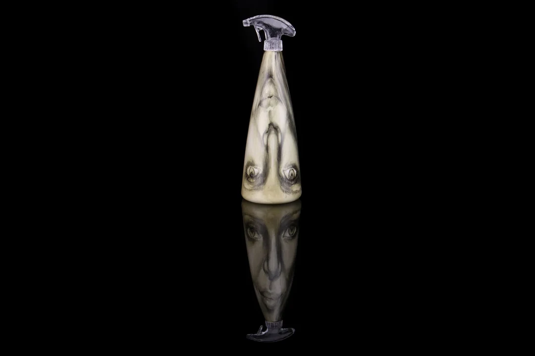 A tall conical glass spray bottle with a woman's face painted in dark enamels on its pale surface, photographed against black with its reflection below.