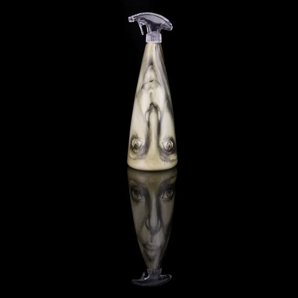 A tall conical glass spray bottle with a woman's face painted in dark enamels on its pale surface, photographed against black with its reflection below.