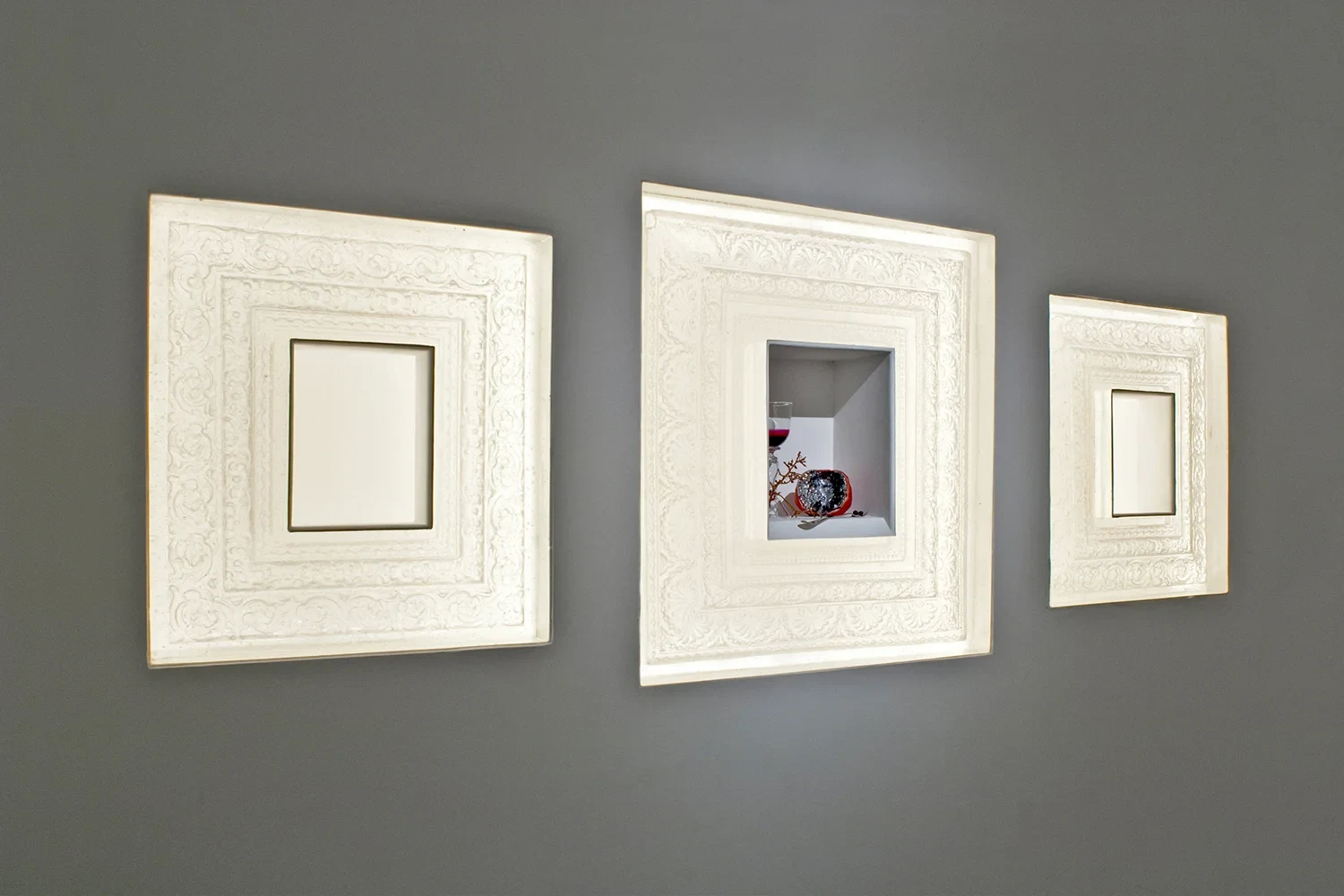Three ornate white cast-glass frames on a gray wall; the middle frame holds a pomegranate, a wine glass and a withered stem.