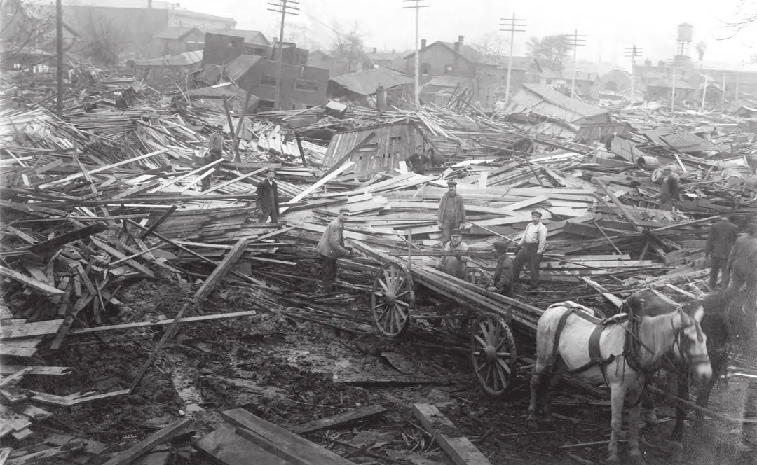 Workers with a horse-drawn wagon salvage lumber among huge piles of flood wreckage, buildings behind. Image from A Flood of Memories (Miami Conservancy District, 2013).