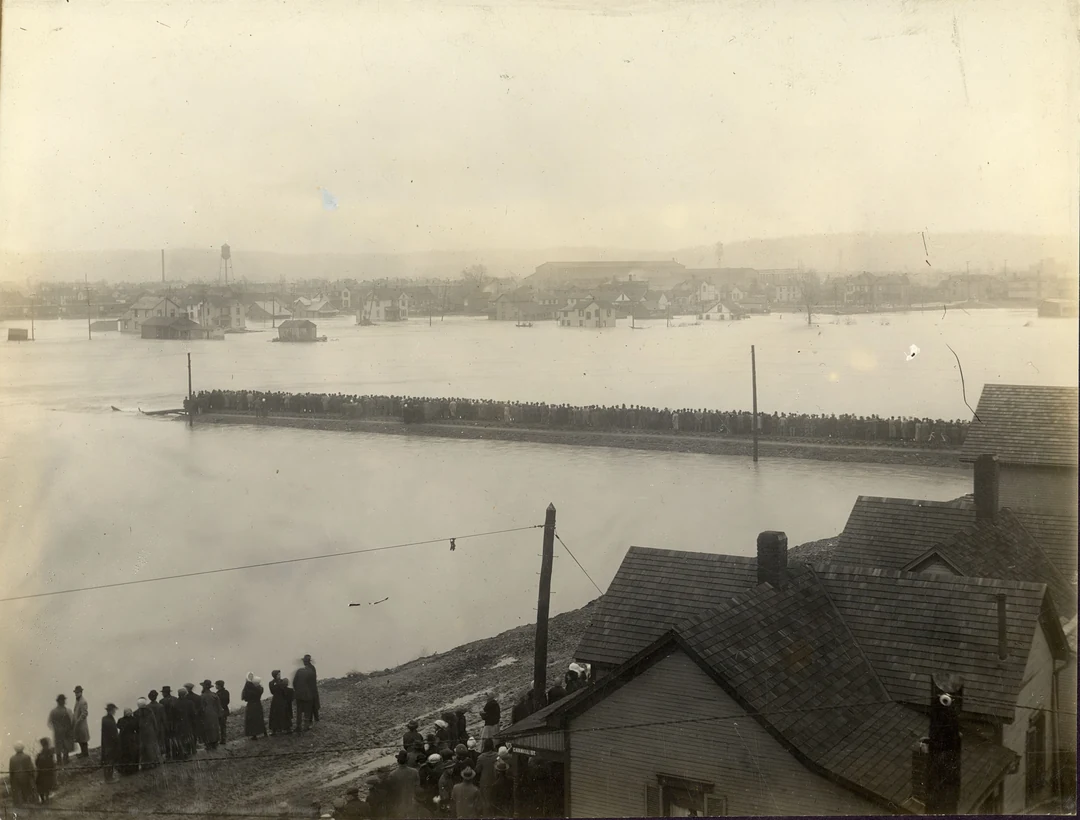 The flooded Great Miami River at Dayton, Ohio, 1913, seen from a hill: a long embankment crosses the water, rooftops in the lower right, a crowd watching at lower left. Image from Dayton Metro Library.