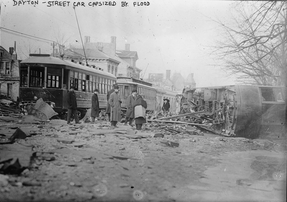 Black-and-white 1913 photograph of a streetcar tipped on its side by floodwaters in Dayton, Ohio, with onlookers standing among debris; handwritten caption at top reads 'Dayton - street car capsized by flood'.
