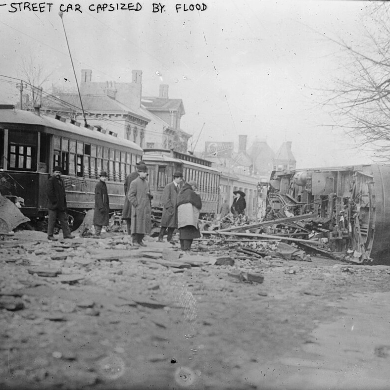 Black-and-white 1913 photograph of a streetcar tipped on its side by floodwaters in Dayton, Ohio, with onlookers standing among debris; handwritten caption at top reads 'Dayton - street car capsized by flood'.