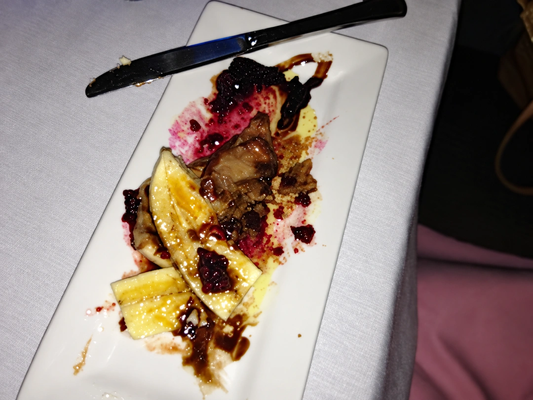 Half-eaten dessert with banana and berries on a rectangular white plate, a knife above it on a white tablecloth. Photograph by Louis Zoellar Bickett. Image from the AEQAI archive. (Digitally enhanced. May contain discrepancies from the original artwork.)